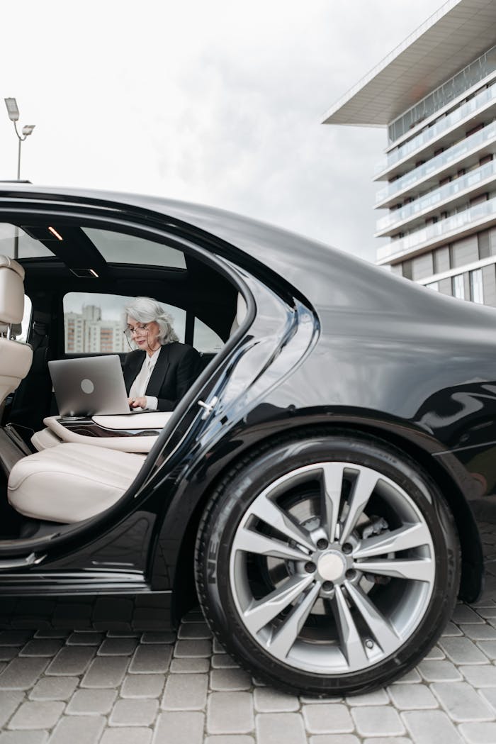 Senior businesswoman using a laptop inside a black car parked outdoors.