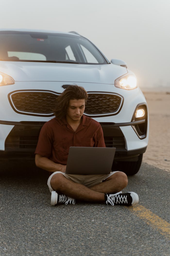 Man sitting with a laptop in front of a car on a desert road at dusk, enjoying a digital nomad lifestyle.